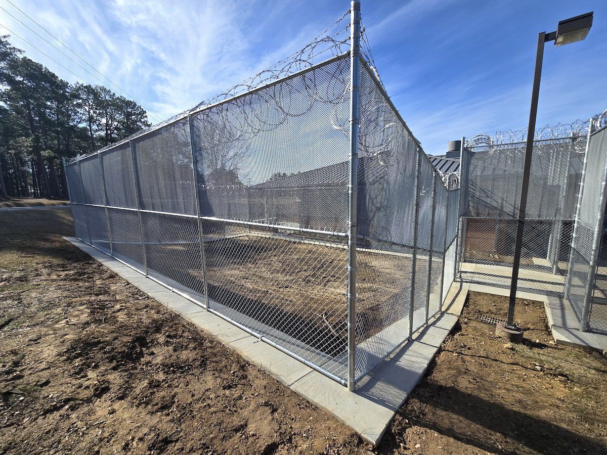 A chain-link security fence with barbed wire topping stands on a concrete foundation in a dirt lot under a blue sky.