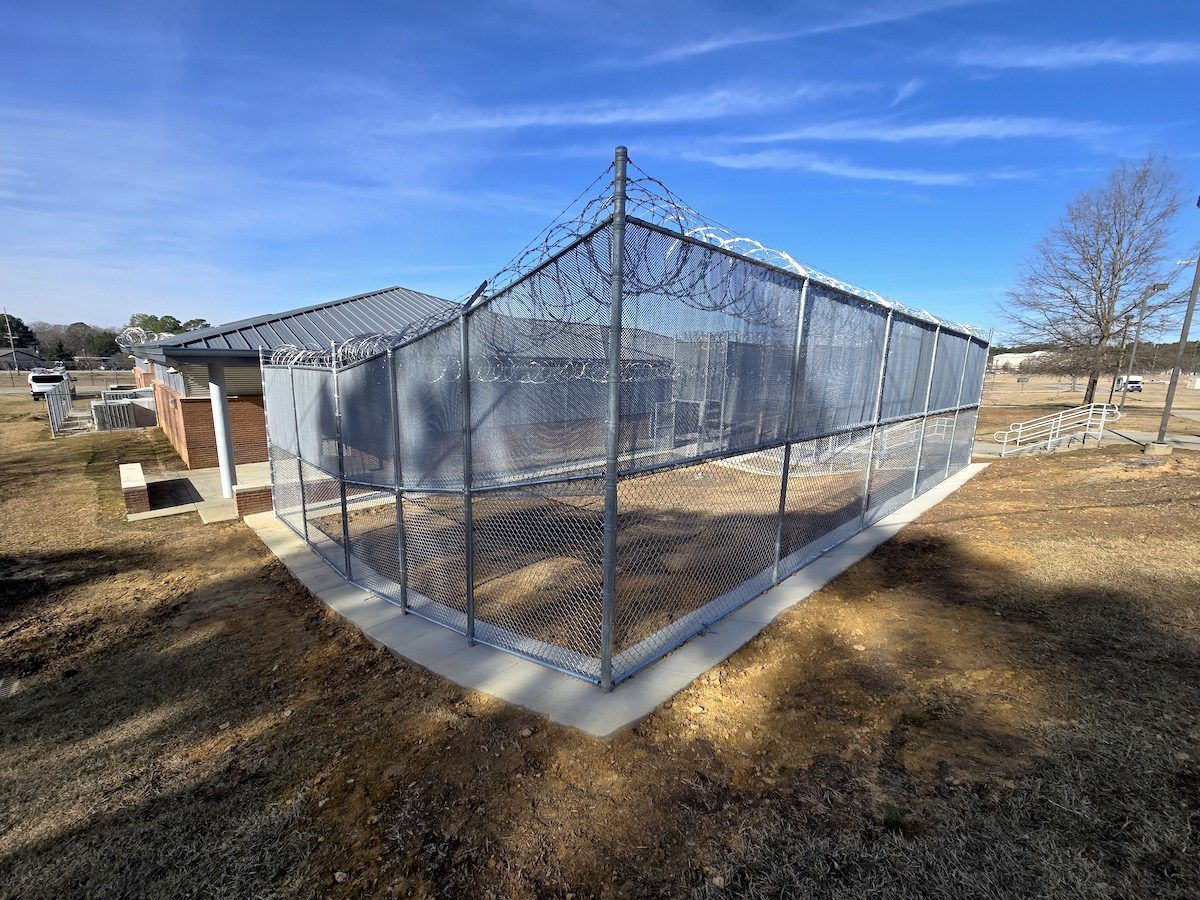 A fenced, concrete-pad enclosure with barbed wire, located outdoors in a grassy area near a building under a blue sky.