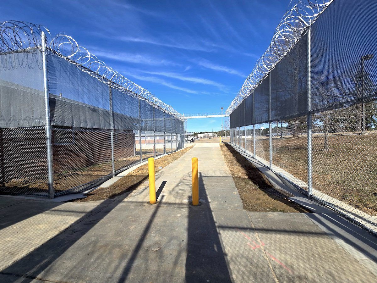 A narrow, paved walkway between two high chain-link fences topped with coils of barbed wire under a clear blue sky.