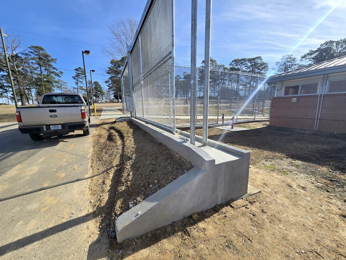 A tall metal fence mounted on a concrete base, adjacent to a parked light-colored pickup truck on an asphalt road.