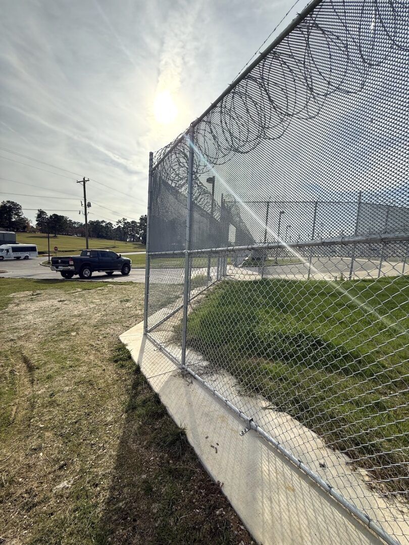 A tall chain-link fence with barbed wire atop a concrete base borders a grassy area, with a truck visible in the distance.