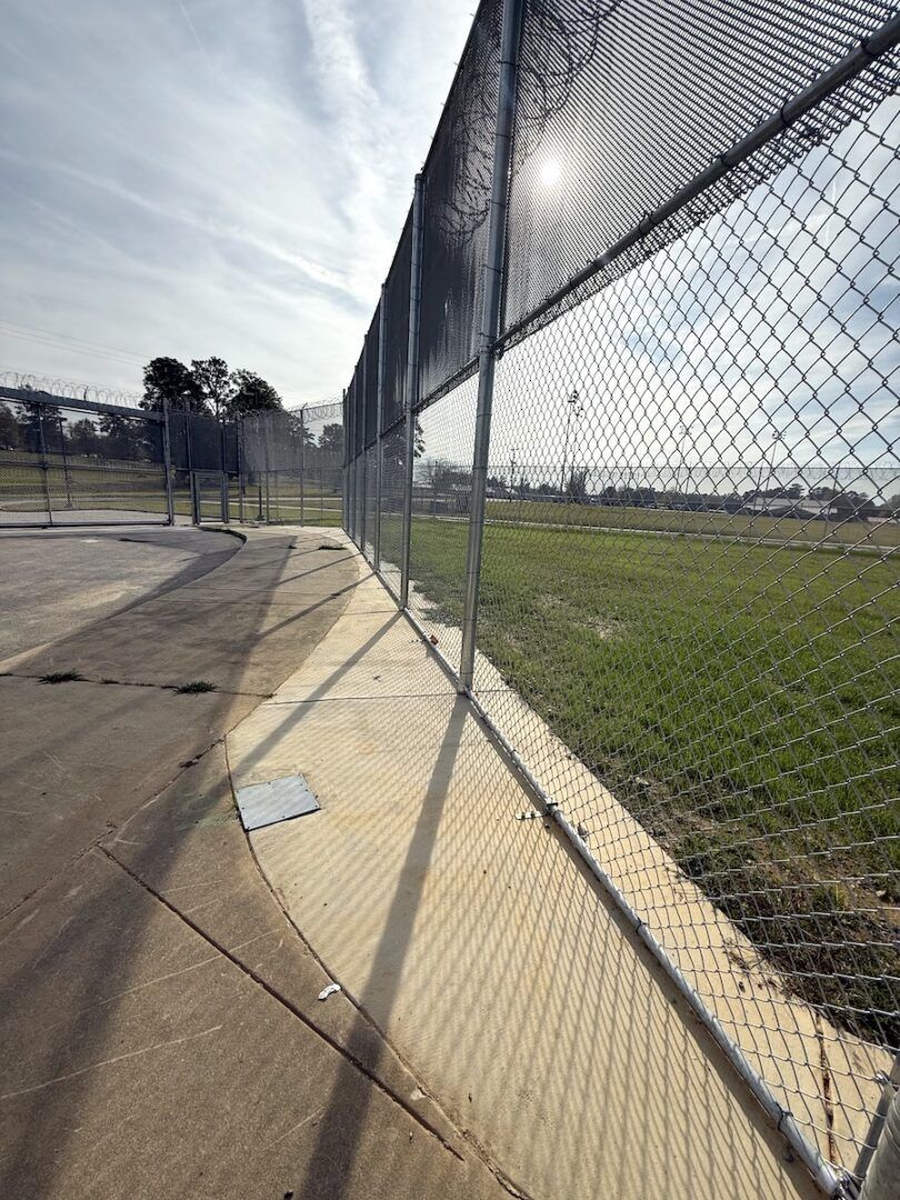 A metal chain-link fence runs alongside a curved sidewalk leading toward a grassy field under a bright sky.