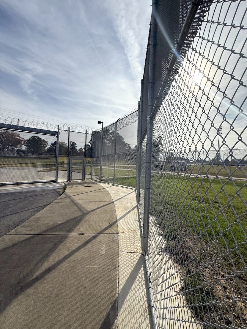 A curved chain-link fence topped with barbed wire lines a concrete path under a bright, sunny sky.