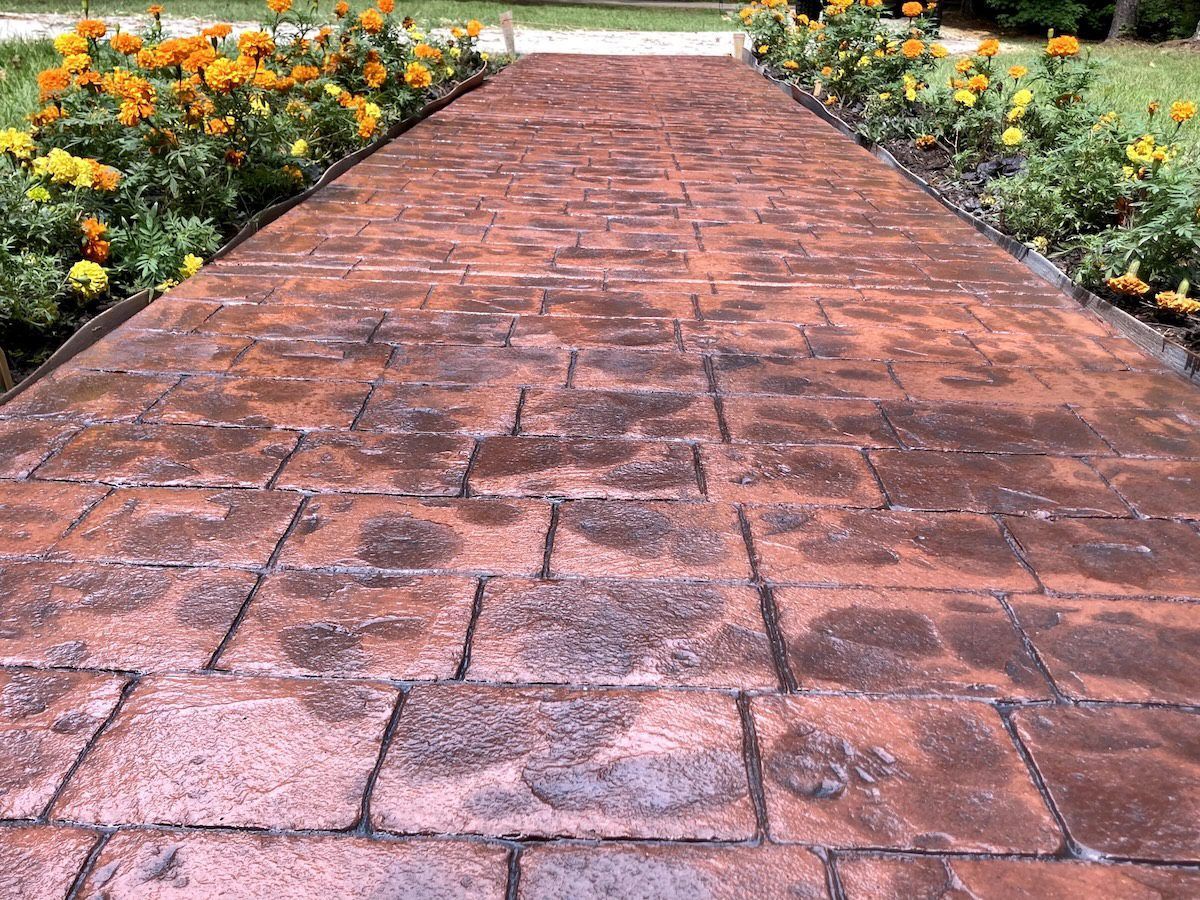 A red, textured, brick-patterned stamped concrete walkway lined with orange marigolds on both sides.