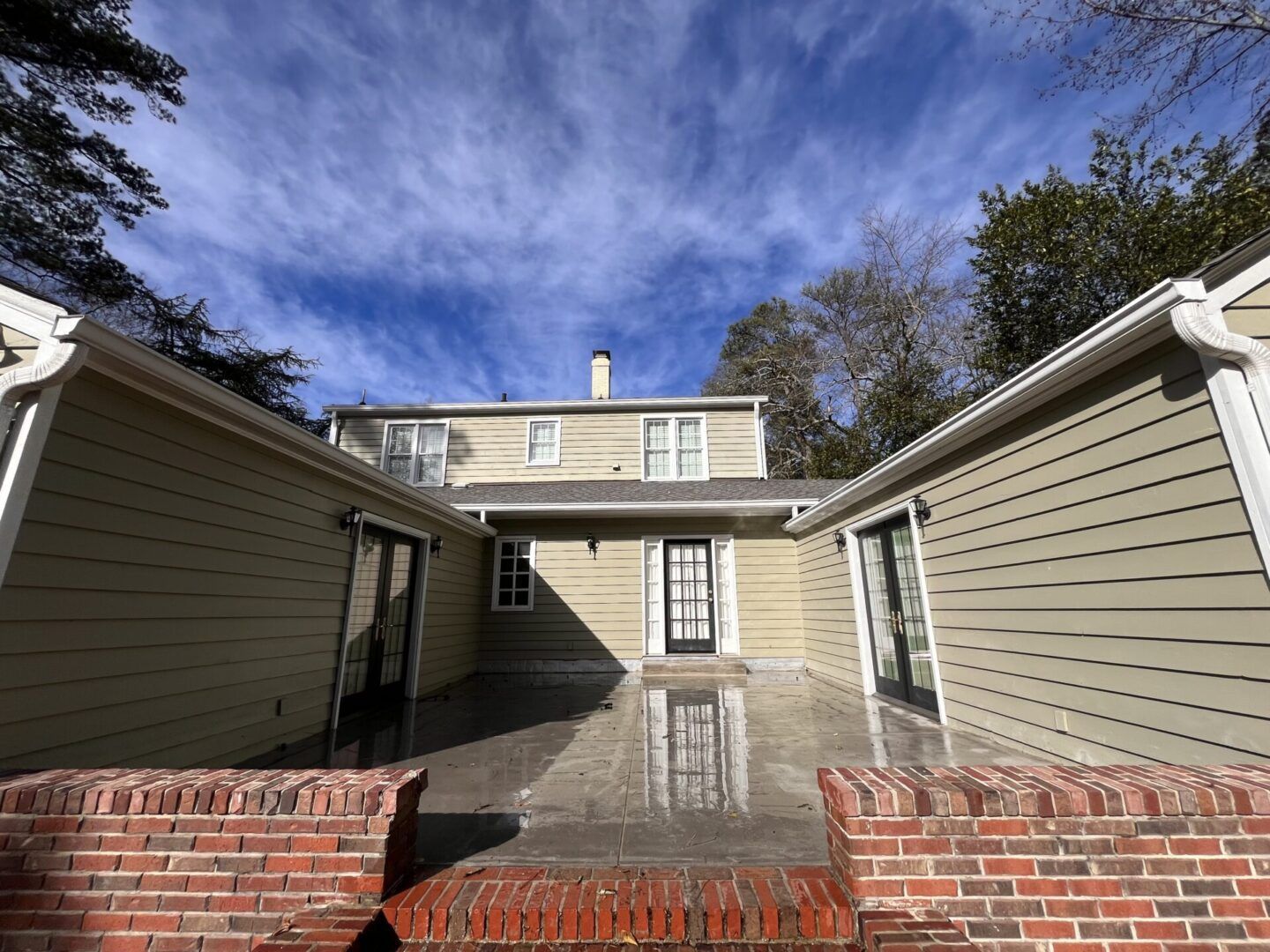 A beige, two-story house with a rear patio, dark-framed doors, and a decorative red brick wall in the foreground.