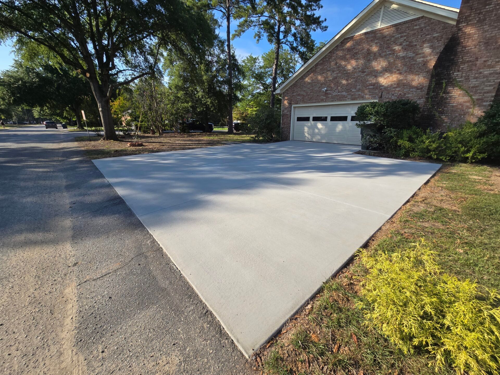 A wide, newly poured concrete driveway leads to a brick house garage in a sunny, tree-lined residential neighborhood.