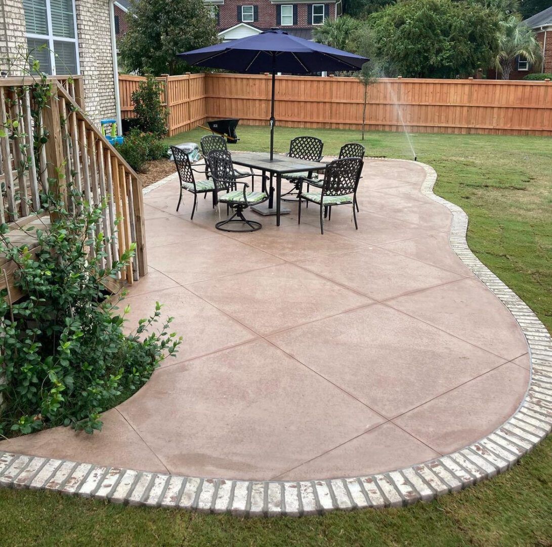 A backyard patio featuring a concrete surface with diagonal scoring and a light brick border, set with a table and chairs.