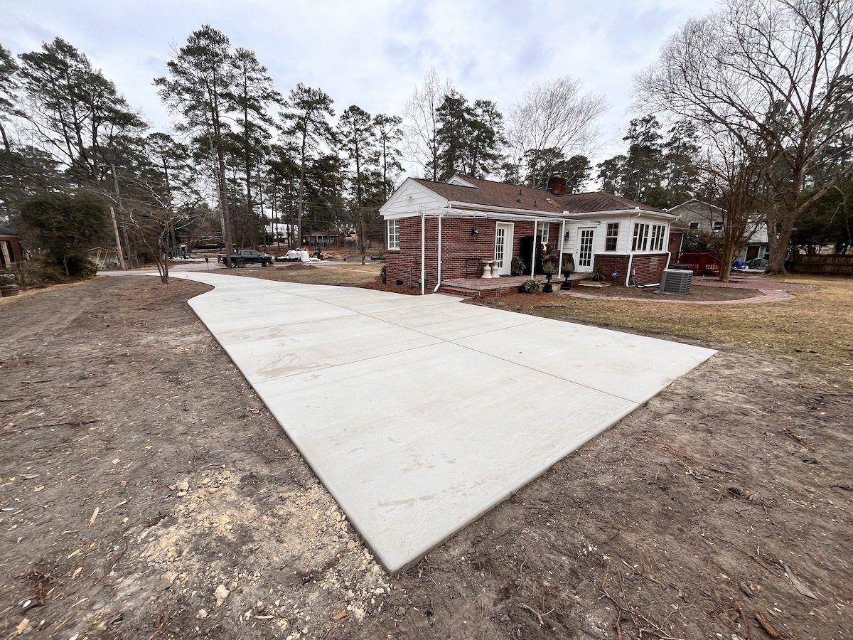 A wide, newly poured concrete driveway leads to the side entrance of a brick house surrounded by trees on a sunny day.