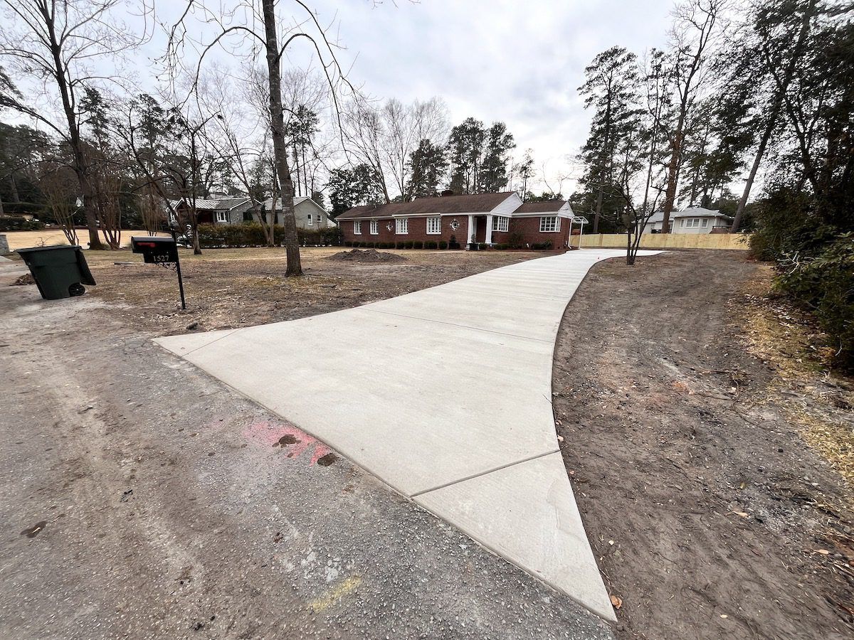 A new, light-gray concrete driveway leads from a gravel road toward a single-story brick house on a suburban lot.