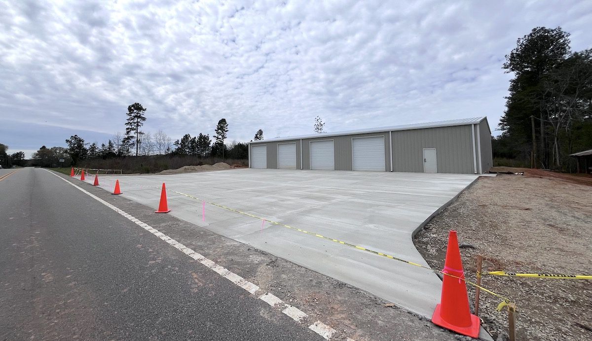 A newly poured concrete parking area leading to a gray metal storage building with four garage doors and safety cones.