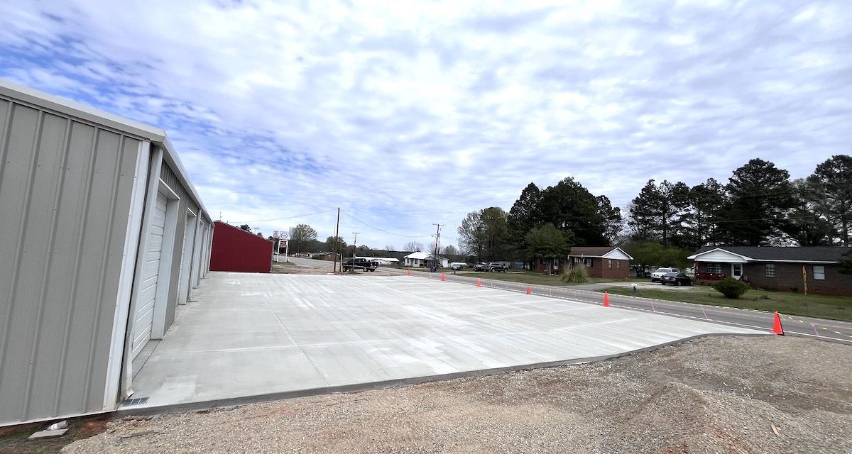A large, freshly poured concrete parking area alongside a light gray storage building under a cloudy blue sky.