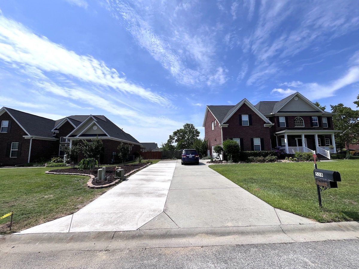 Two brick suburban houses with a shared concrete driveway under a bright blue, cloudy sky.