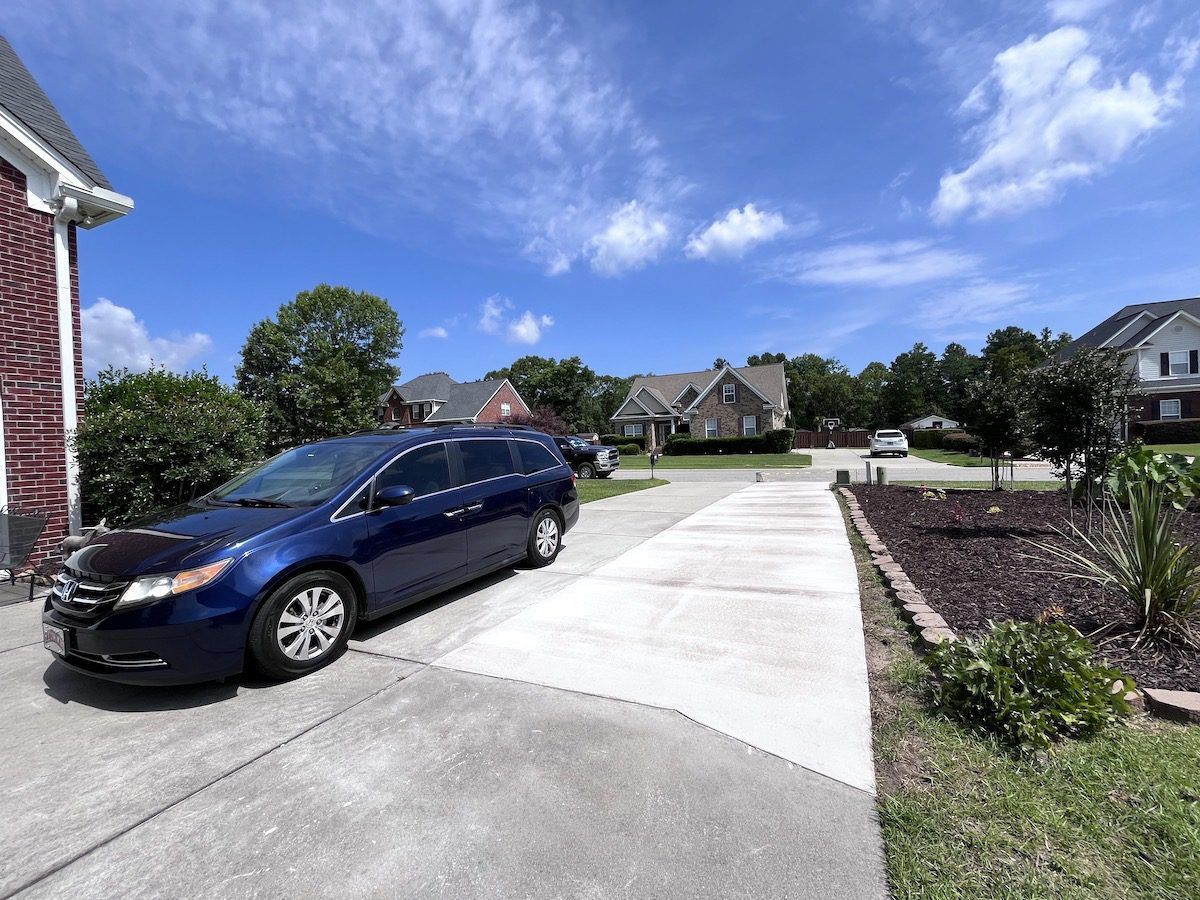 A dark blue minivan parked on a concrete driveway in front of a suburban home under a sunny blue sky.