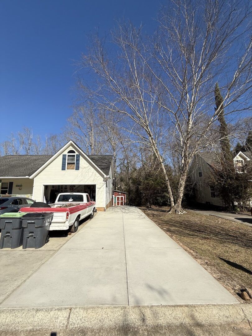 A concrete driveway leads to a garage and house, with a truck parked on the left and trees under a clear blue sky.