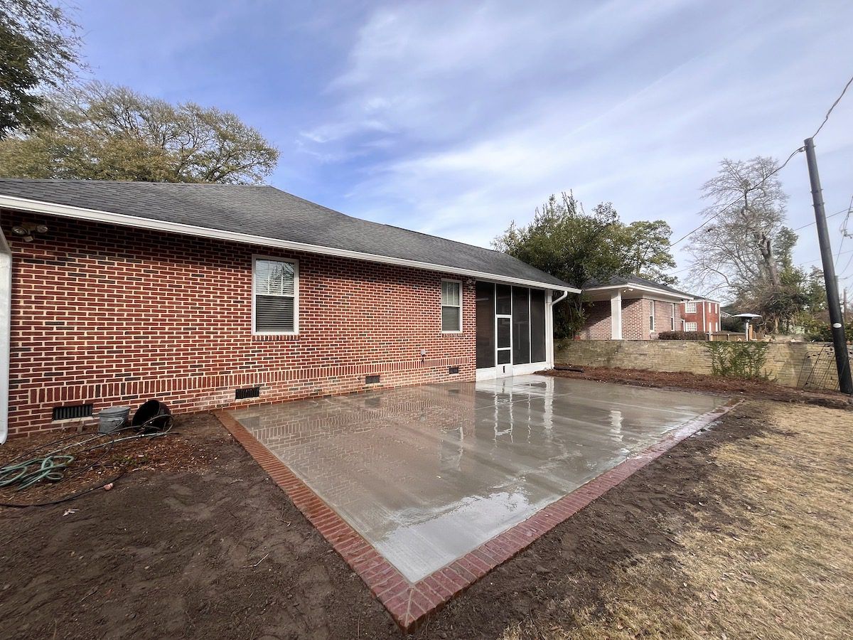 A new rectangular concrete patio with a brick border sits in the backyard of a brick house under a blue sky.
