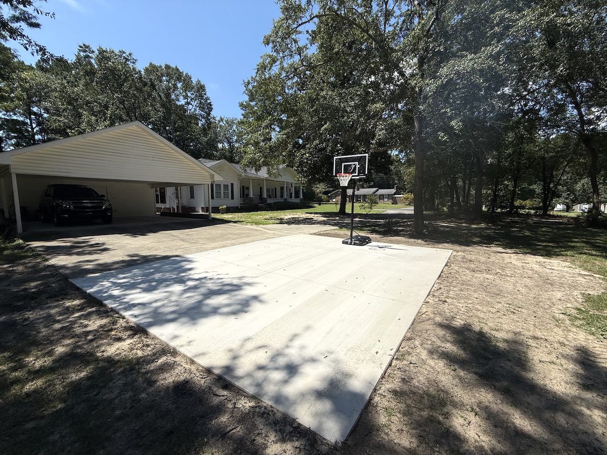 A white car parked under a carport next to a concrete basketball court in a wooded yard.
