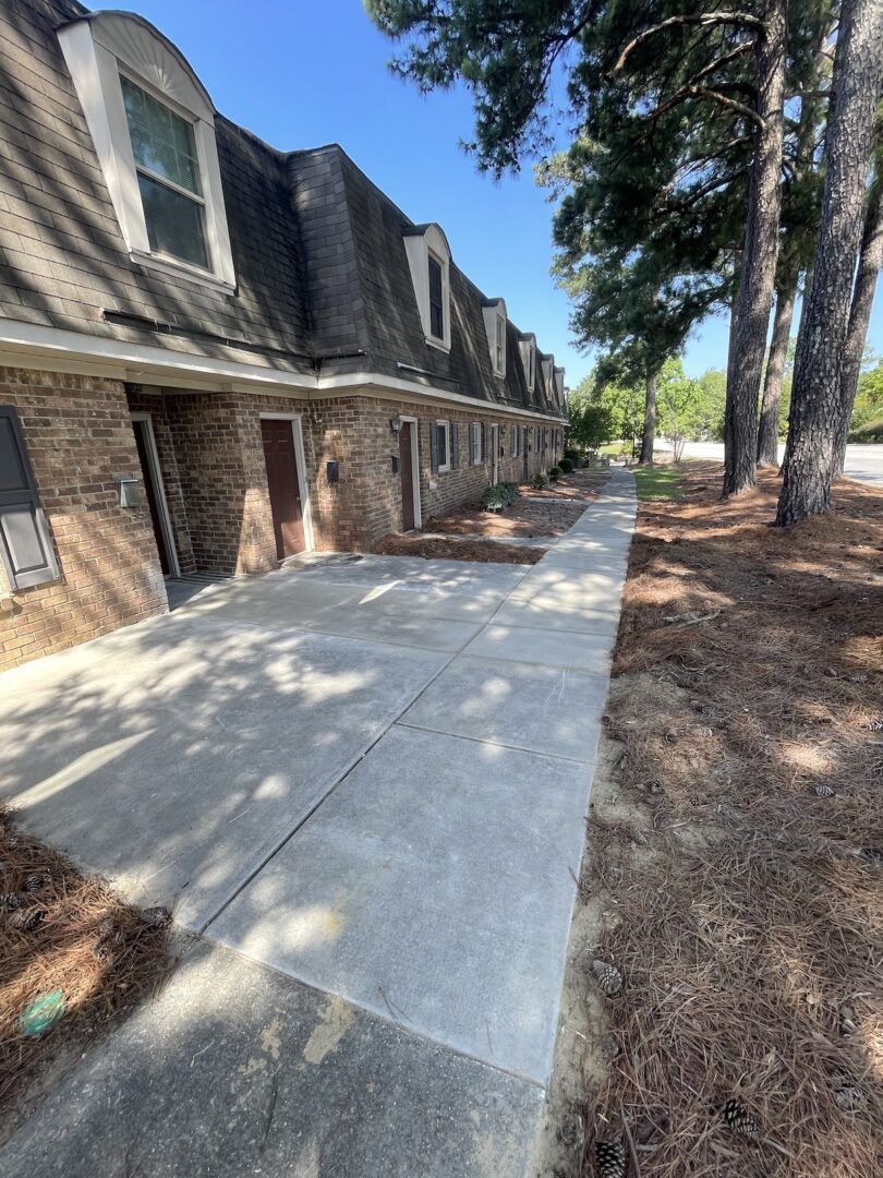 A long, brick apartment building with a gray roof and concrete walkway sits beside a dirt area with several tall trees.