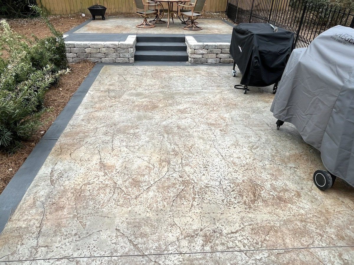 A split-level backyard patio with stamped concrete, stone steps, a covered grill, and outdoor furniture.