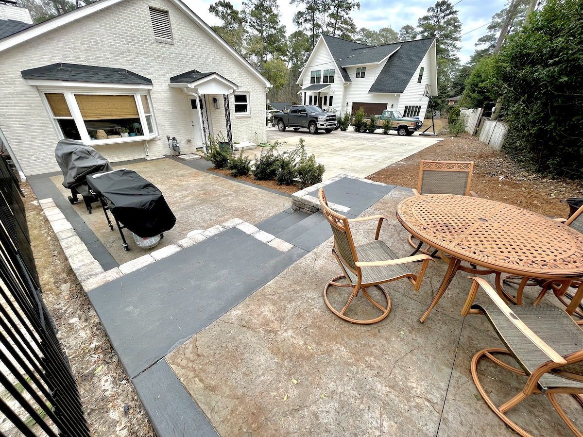 A patio with wicker furniture, a grill, and gray pavers in front of a white house with a driveway and another home nearby.