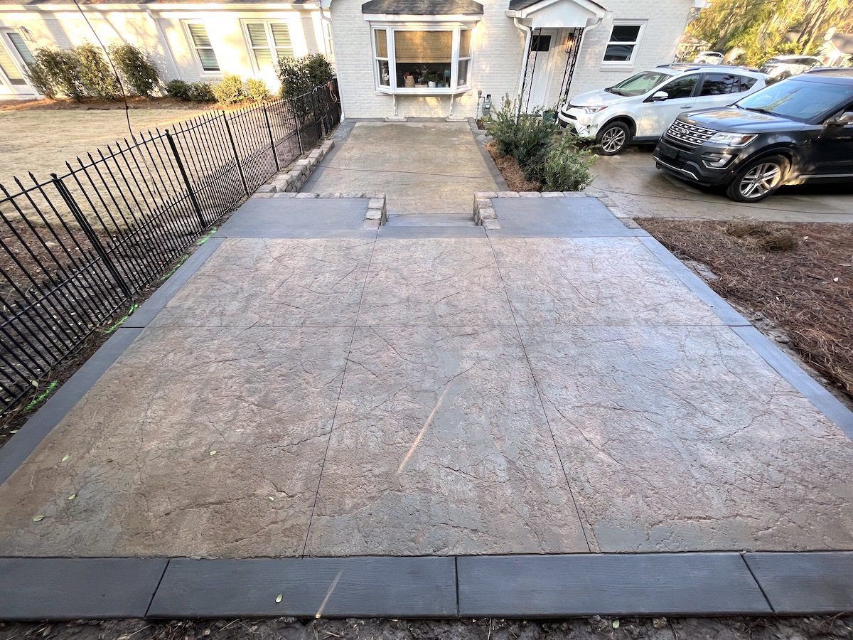 Newly poured concrete patio featuring a light, textured center bordered by smooth, dark gray pavers in a suburban yard.