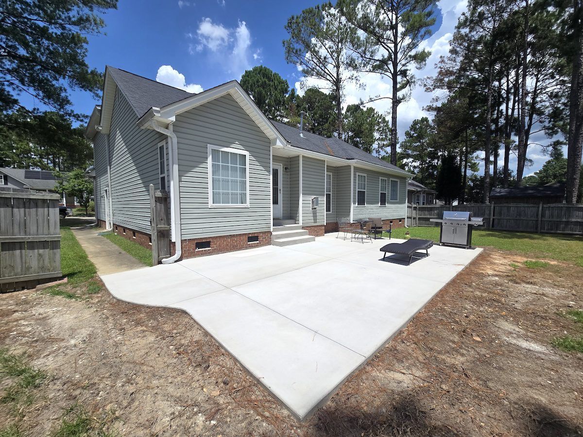 Light gray house with a brick foundation and a spacious concrete patio in a backyard with trees under a blue sky.