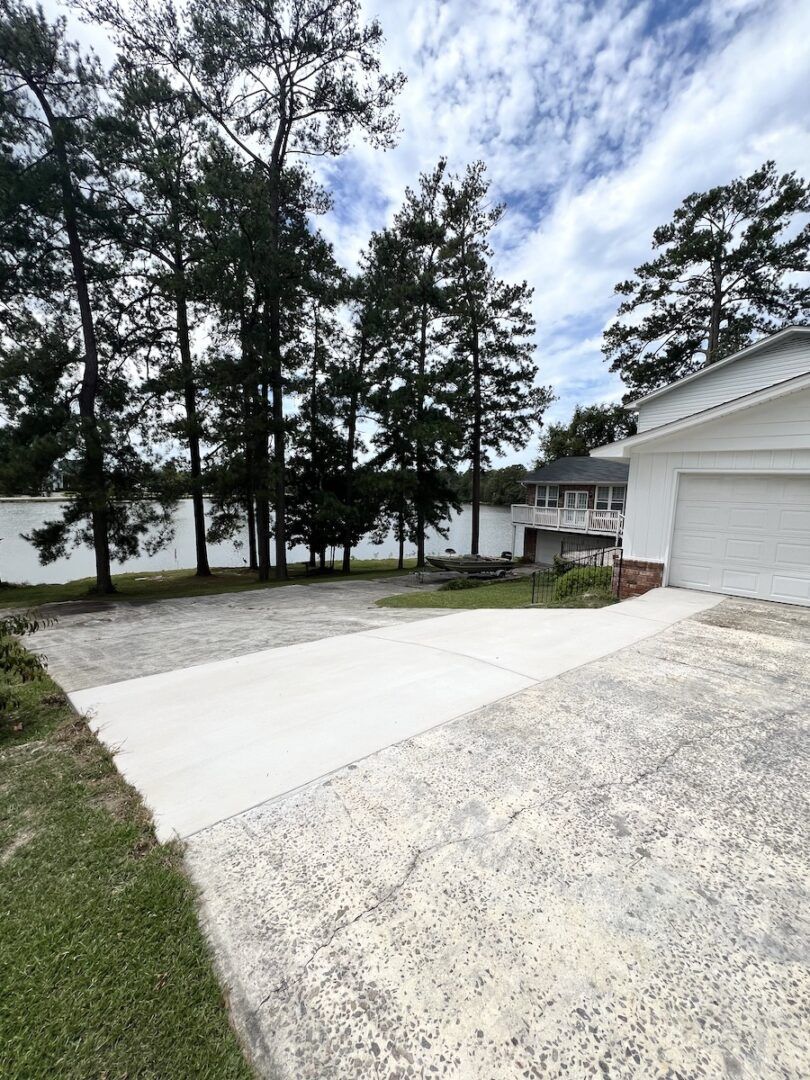 A wide gravel and concrete driveway leads toward a house with a white garage, surrounded by tall pine trees.
