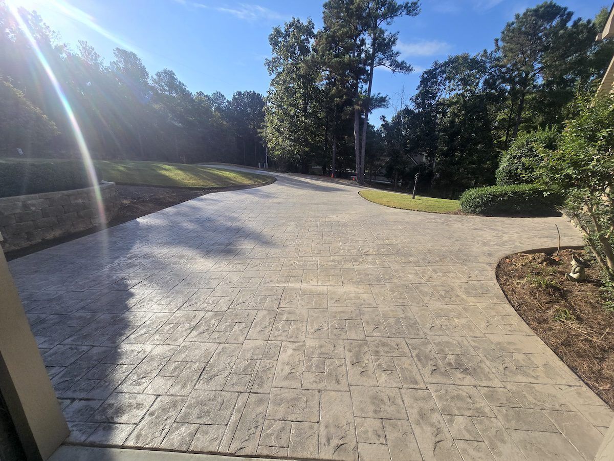 A stamped concrete driveway leads toward a tree-lined yard under a bright, sunny sky.