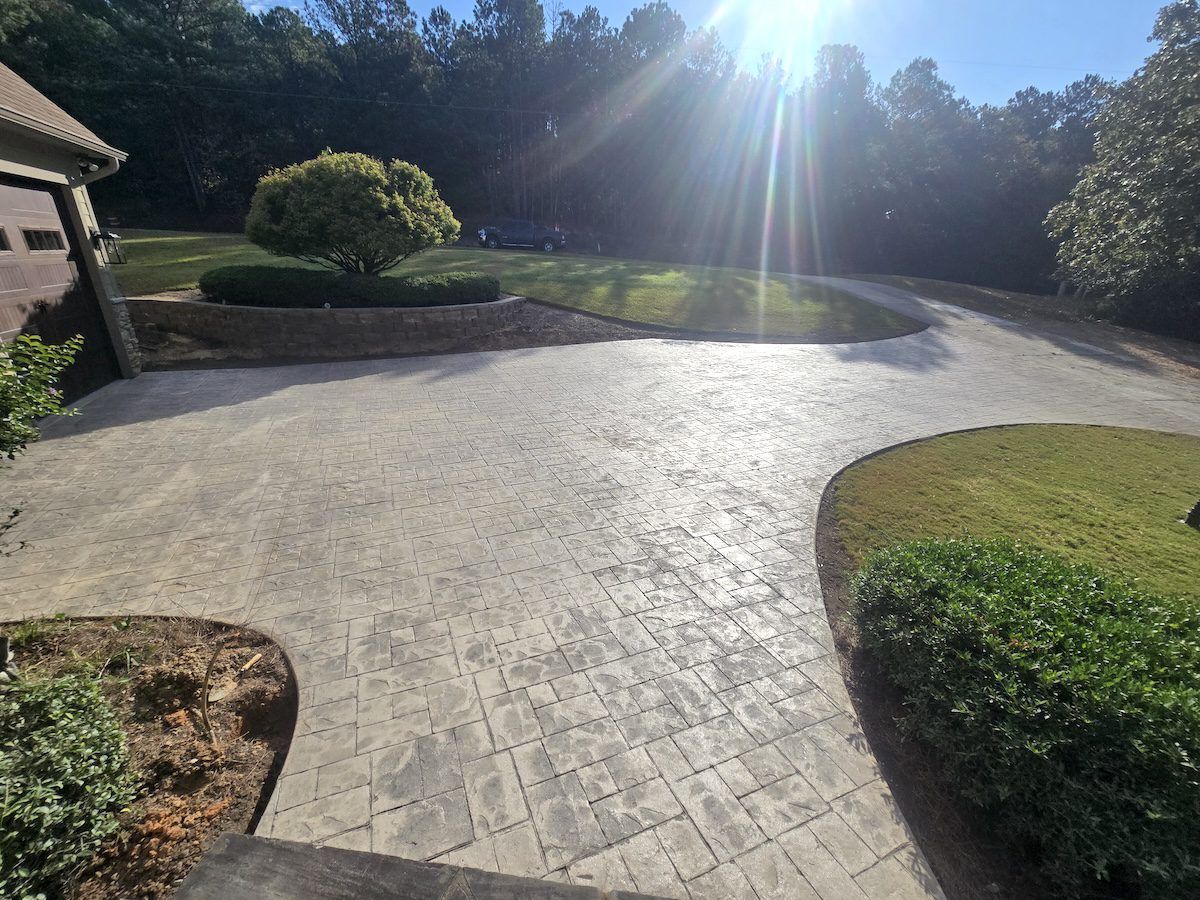 A bright, sunny outdoor view of a stone-paver driveway leading toward a house, surrounded by green lawns and trees.