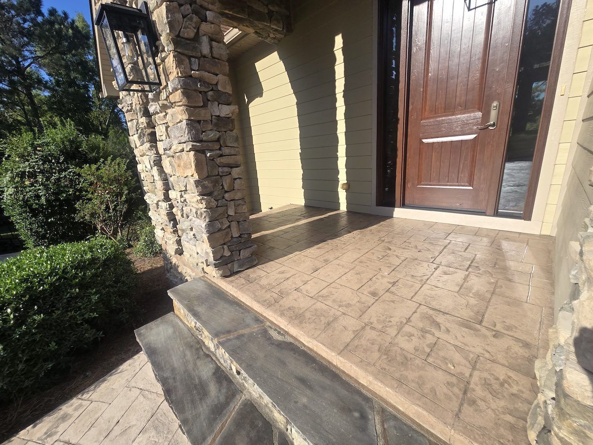 A front porch with tan stamped concrete flooring, dark stone steps, and a wooden front door, set against a stone pillar.