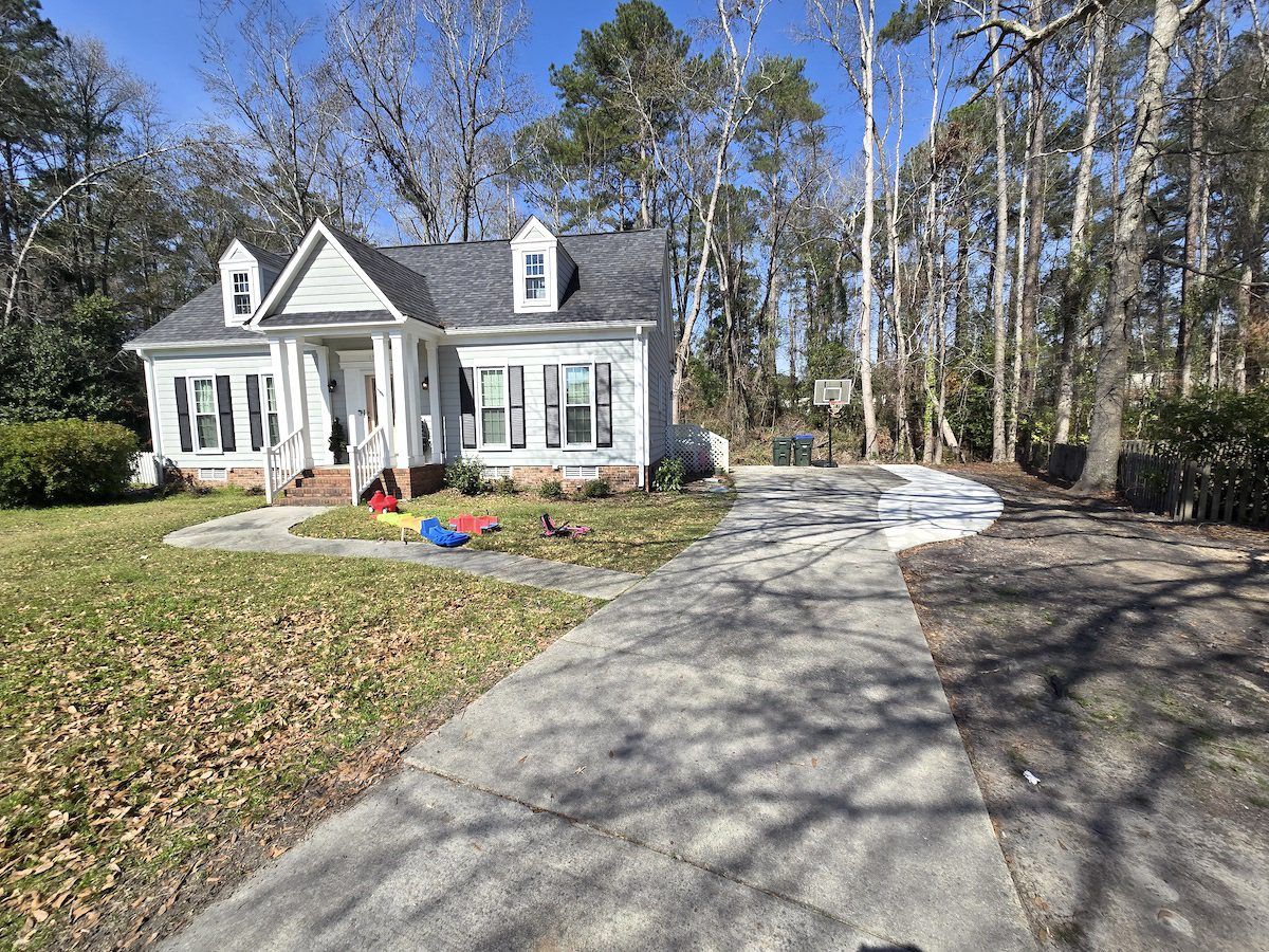 A light-colored two-story house with a long concrete driveway, front lawn, and a backdrop of tall pine trees.