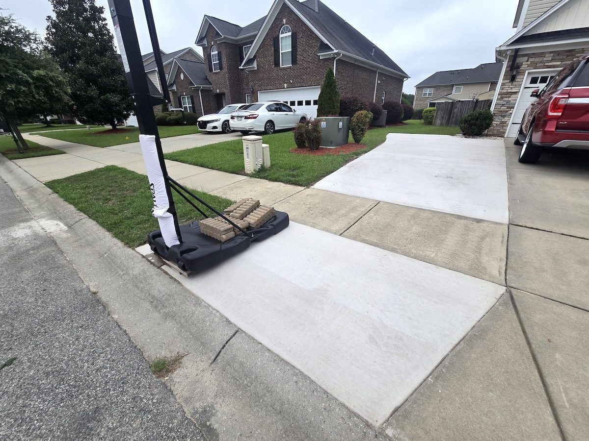 A portable basketball hoop sits on a newly poured concrete driveway patch next to a neighborhood street and house.