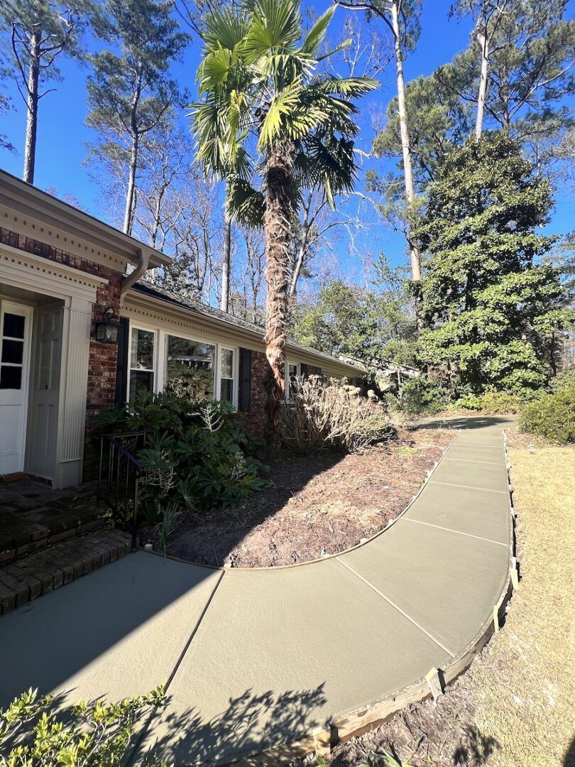 A brick house exterior with a curved concrete sidewalk, surrounded by trees and a tall palm tree under a blue sky.