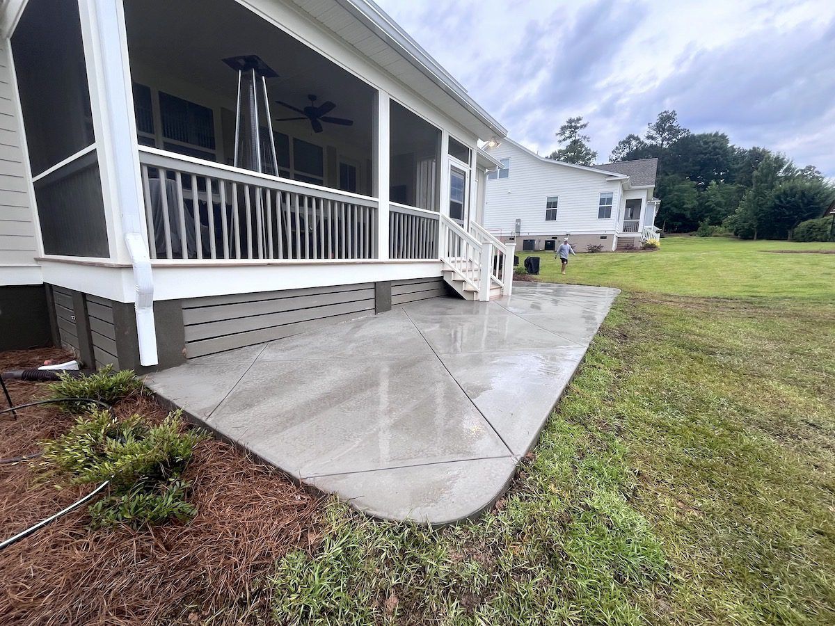 A ground-level view of a newly poured concrete patio outside a house with a screened-in porch and stairs.
