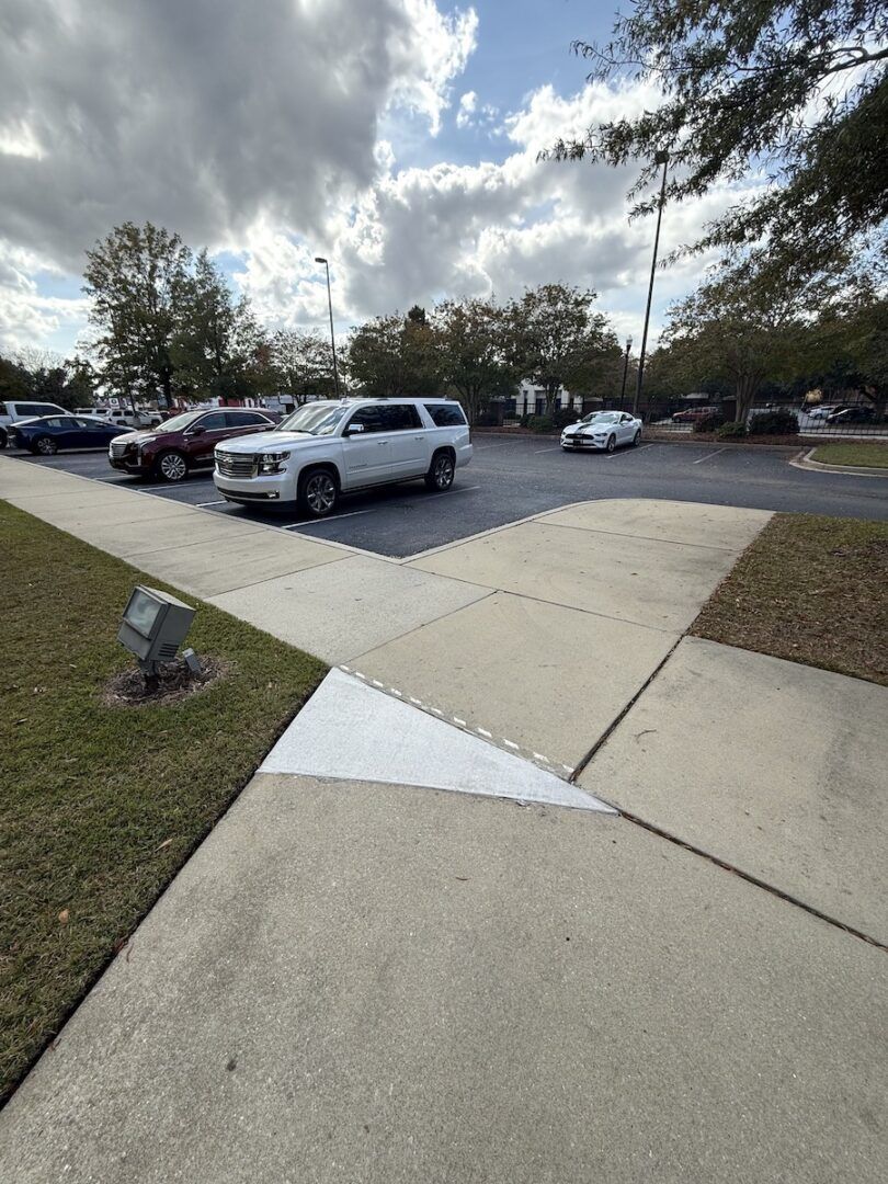 A sidewalk leads toward a parking lot with a white SUV and other parked cars under a partly cloudy sky.