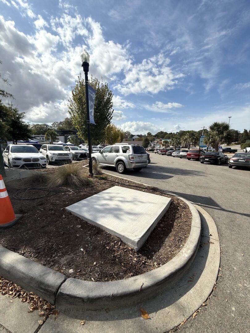 A square concrete pad sits in a landscaped parking island near a lamp post and a row of parked cars under a blue sky.