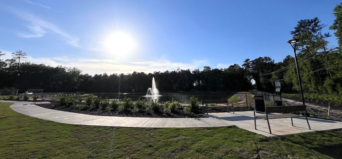 A sunny park scene with a paved path curving around a central pond featuring a fountain, set against a line of trees.