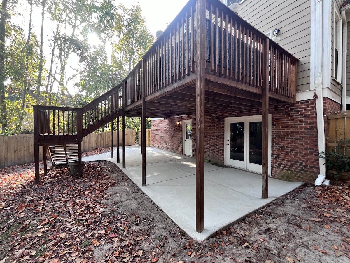 A wooden deck with a staircase overlooks a concrete patio against a brick and siding house exterior on a sunny day.