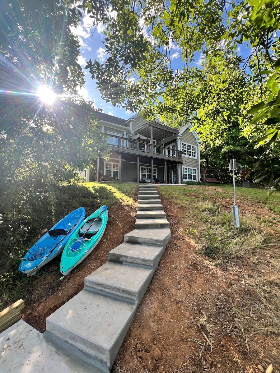A concrete stairway leads up a grassy slope toward a two-story house, with two kayaks resting on the ground to the left.