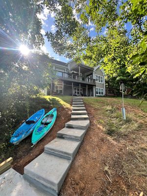 A concrete stairway leads up a grassy slope toward a two-story house, with two kayaks resting on the ground to the left.