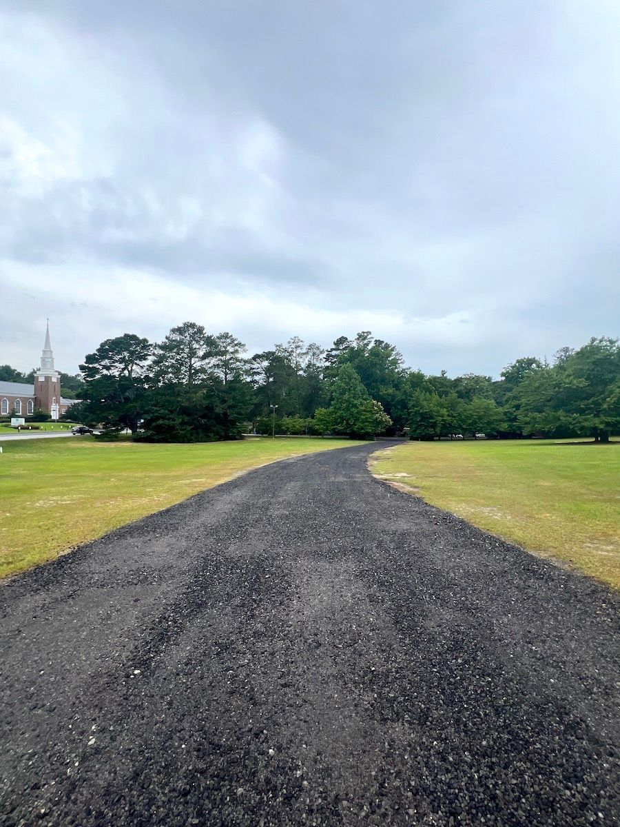A gravel driveway leads through a grassy field toward trees and a church steeple under a cloudy sky.