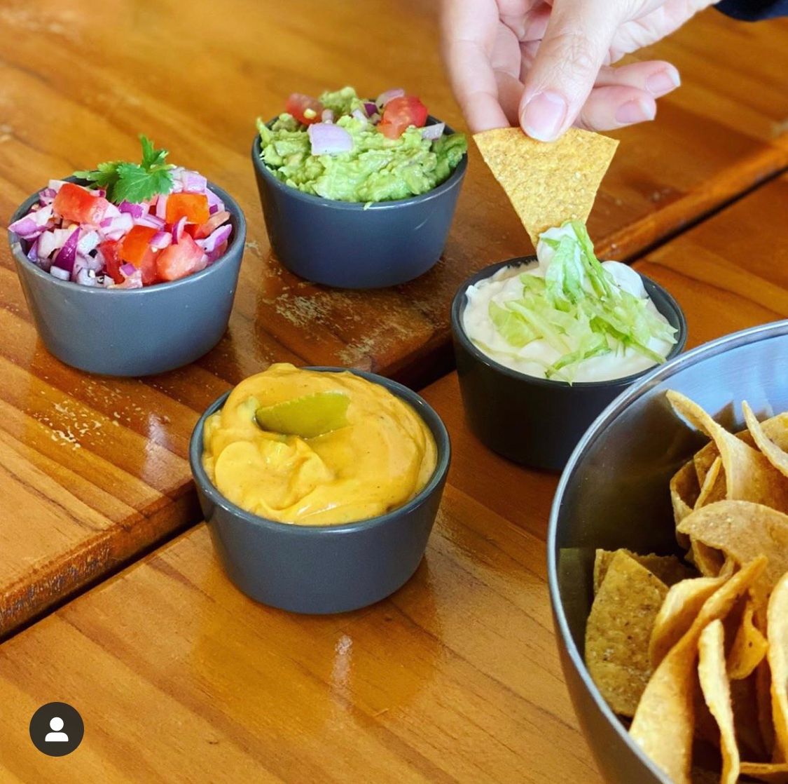 Bowls of dips (guacamole, salsa, cheese sauce, and creamy dip) with tortilla chips on a wooden table. A hand dips a chip.