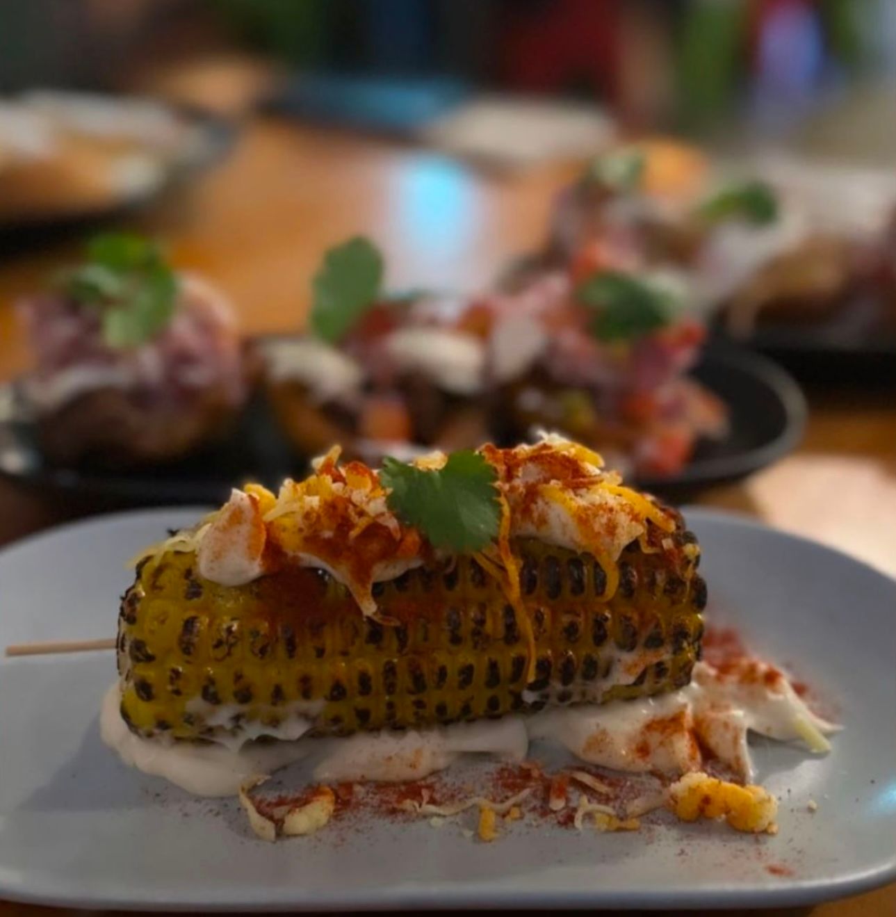 Grilled corn on the cob with toppings, served on a white plate with other tapas.