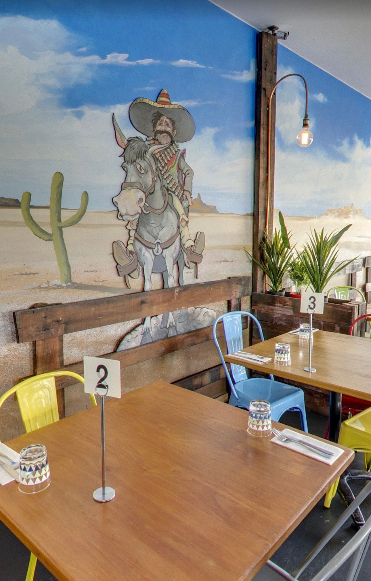 Restaurant interior with tables and chairs, a mural of a man on a donkey, and desert scenery.