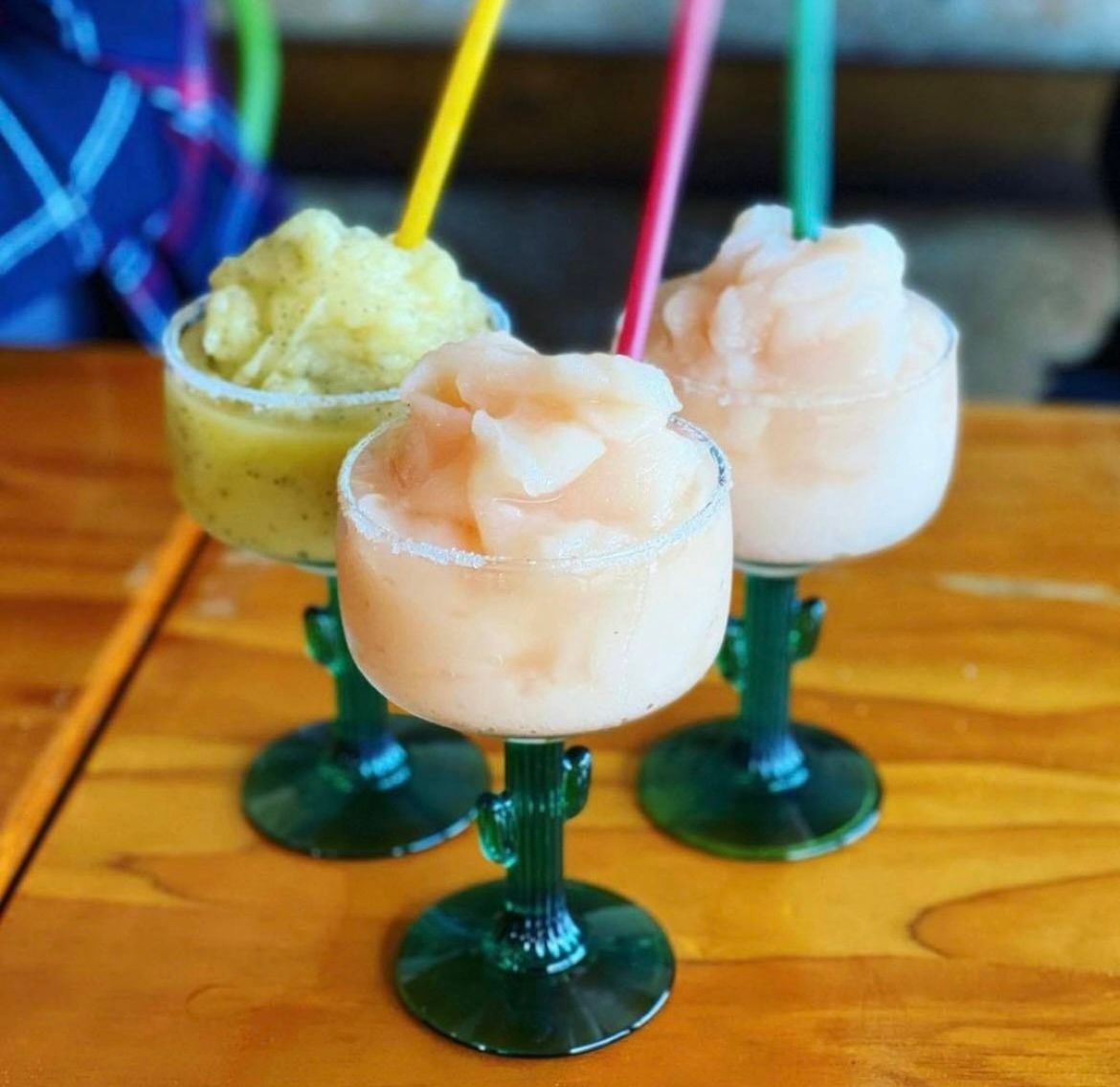 Three fruity frozen margaritas in cactus-shaped green glasses, with straws, on a wooden table.