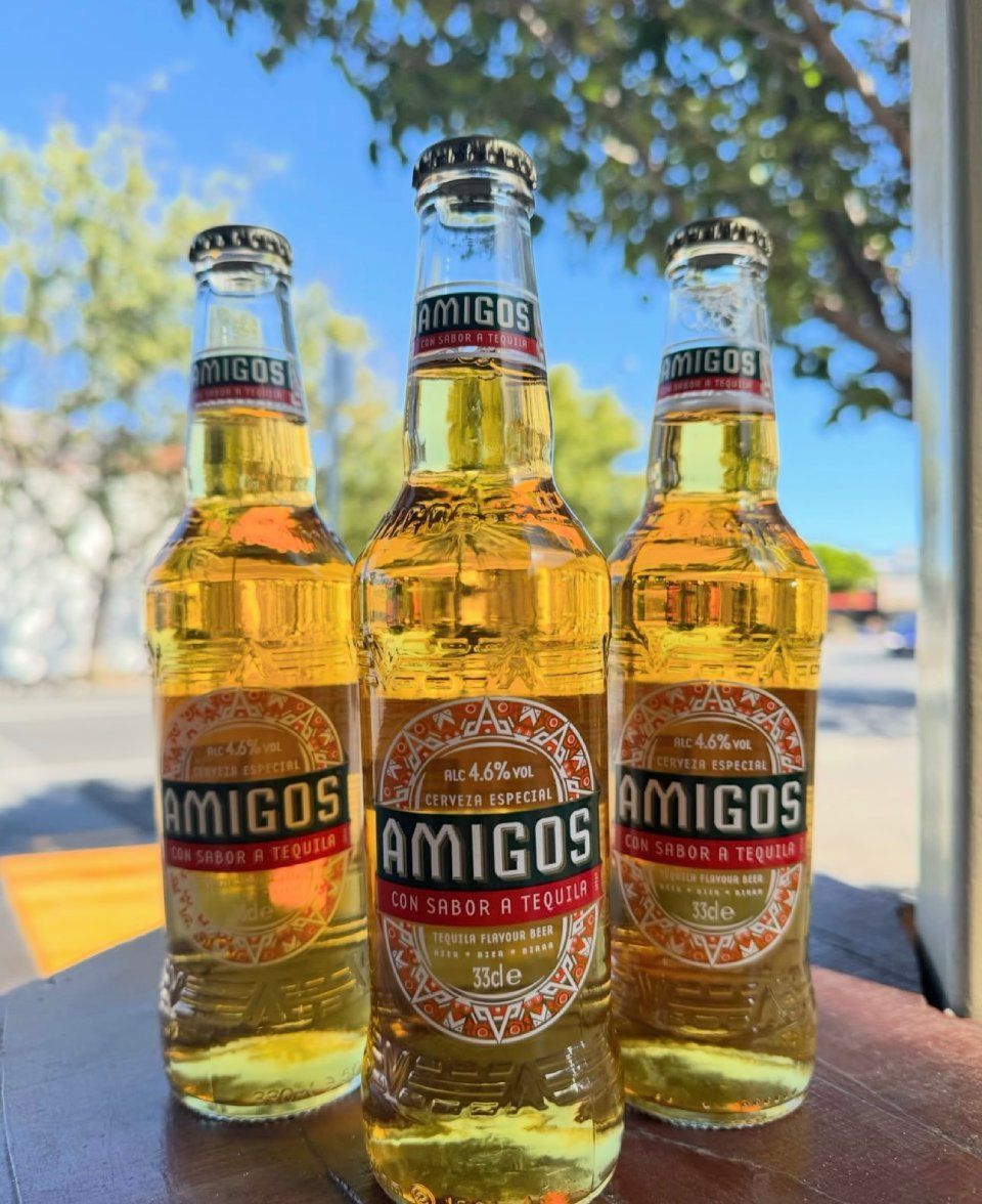 Three bottles of Amigos beer, golden liquid with red and gold labels, on a table outdoors.