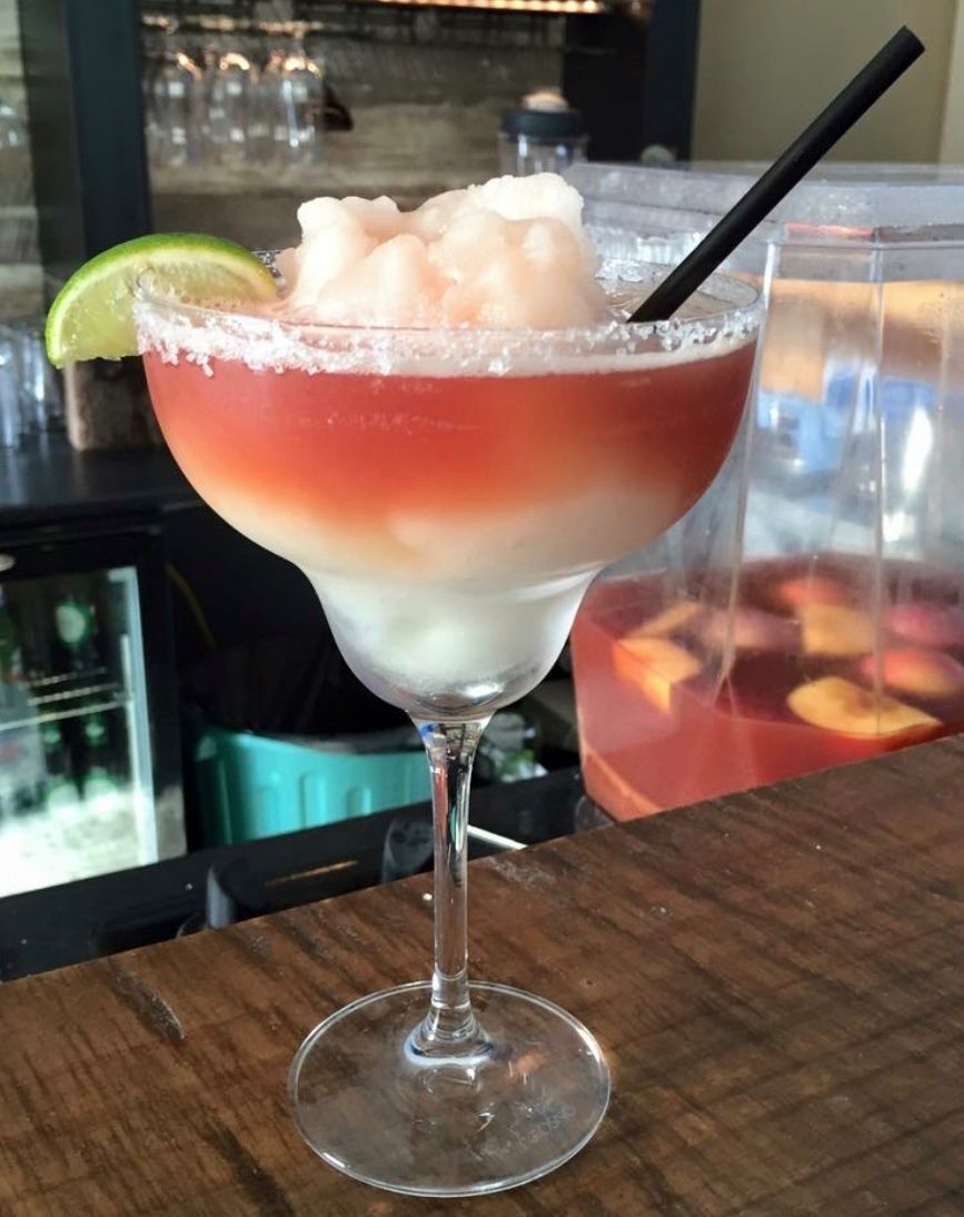 A layered cocktail in a stemmed glass, rimmed with salt, topped with ice, lime slice, and black straw.