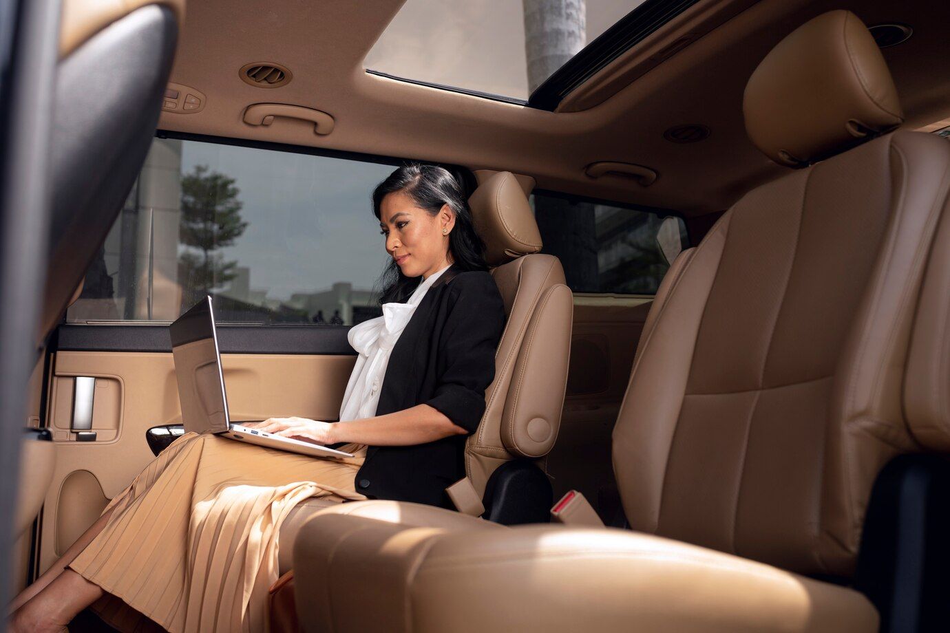 A woman is sitting in the back seat of a car using a laptop.