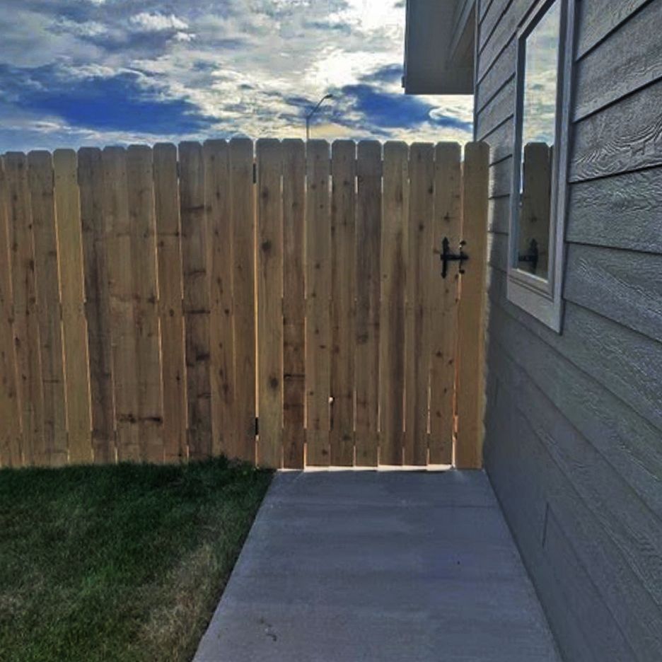 Wooden fence with a gate next to a gray house. A concrete walkway leads to the gate.