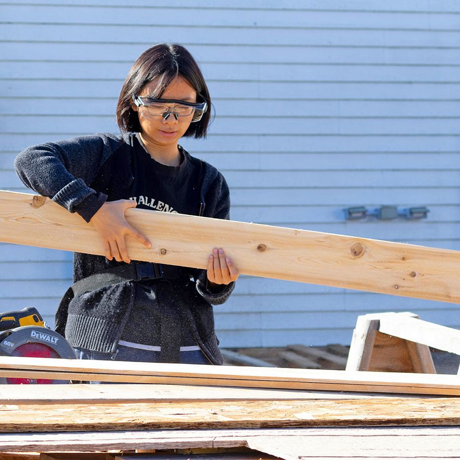 Woman wearing safety glasses, holding a wooden board on a construction site, working on building a structure.
