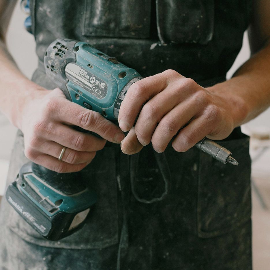 Person in overalls holds a teal and black power drill.